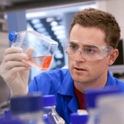 man wearing safety goggles observes a flask of orange liquid